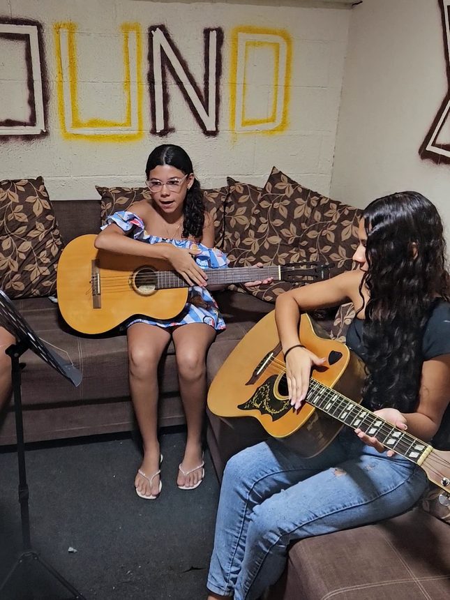 Duas alunas tocando violão lado a lado num sofá