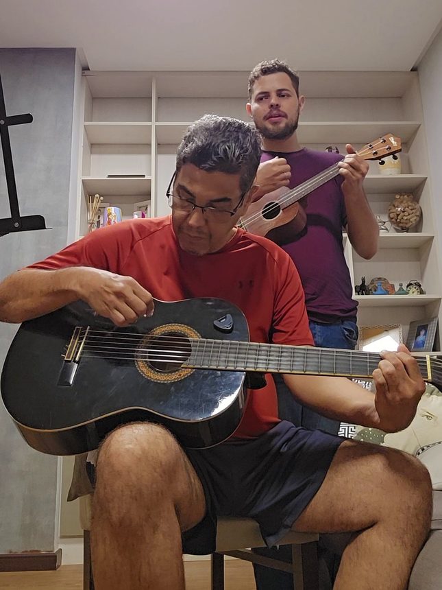 Aluno adulto tocando violão, com o professor ao fundo