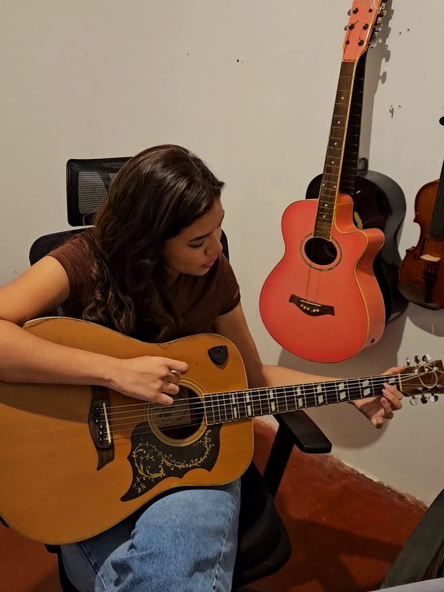 Aluna tocando violão, com violões e violinos pendurados na parede