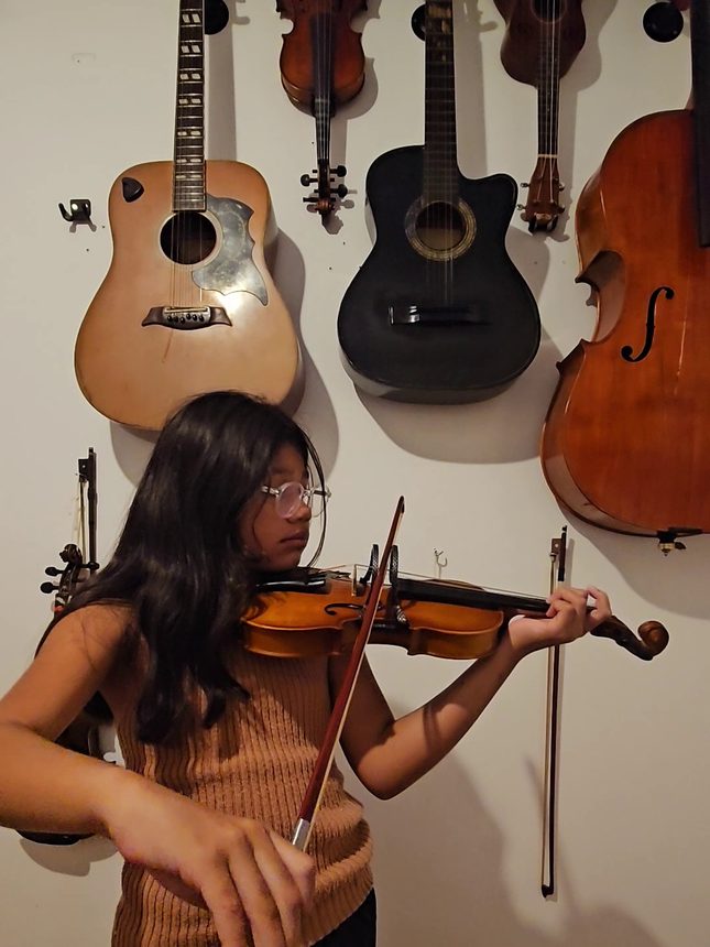 Aluna tocando violino em frente a uma parede de violões